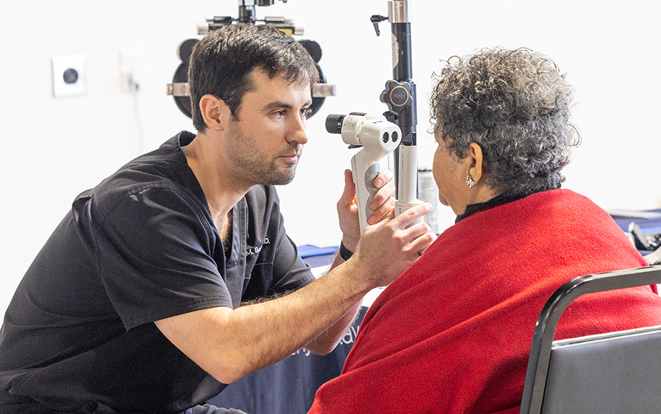 A member of the Optometry Association of Louisiana provides an eye exam to a patient at a VSP Eyes of Hope mobile clinic free vision care outreach event. The optometrist is wearing black scrubs and is examining the health of the eyes of a patient with curly grey hair and a red shawl.