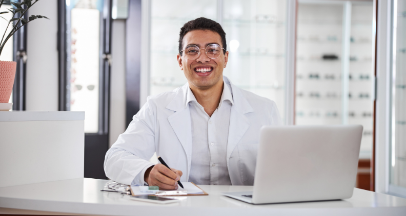 Gentleman wearing glasses and a white button-down shirt and white jacket sitting at a desk with a laptop and a clipboard. He is also holding a pen and smiling directly at the camera.  